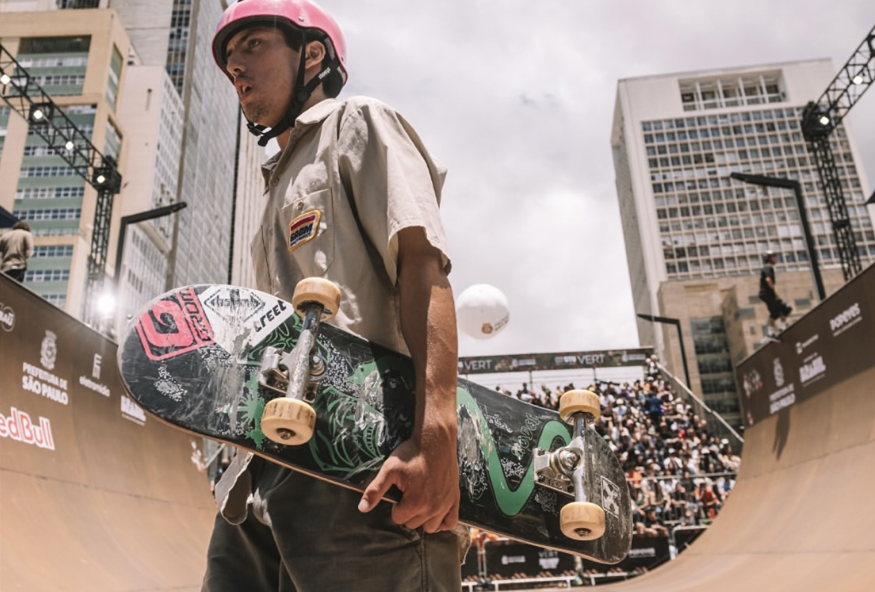 Skateboarder Holding Deck at Competition Park - Thomas Augusto Thomas Augusto holding his skateboard at a skate competition park with a crowd and ramps in the background. - Thomas Augusto